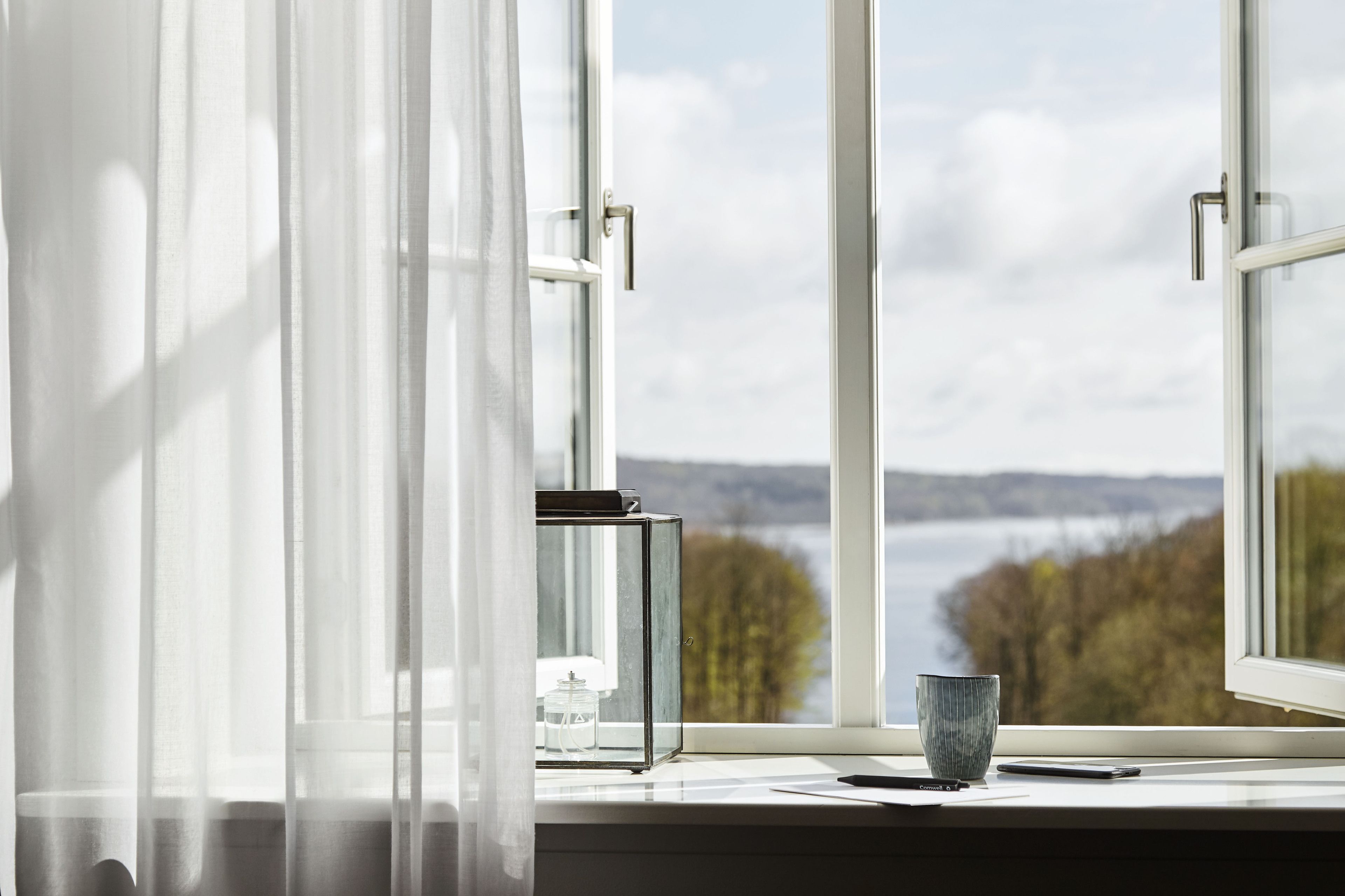 Une fenêtre donnant sur un lac et des arbres, une table en verre avec une tasse et un livre, et des rideaux légers.