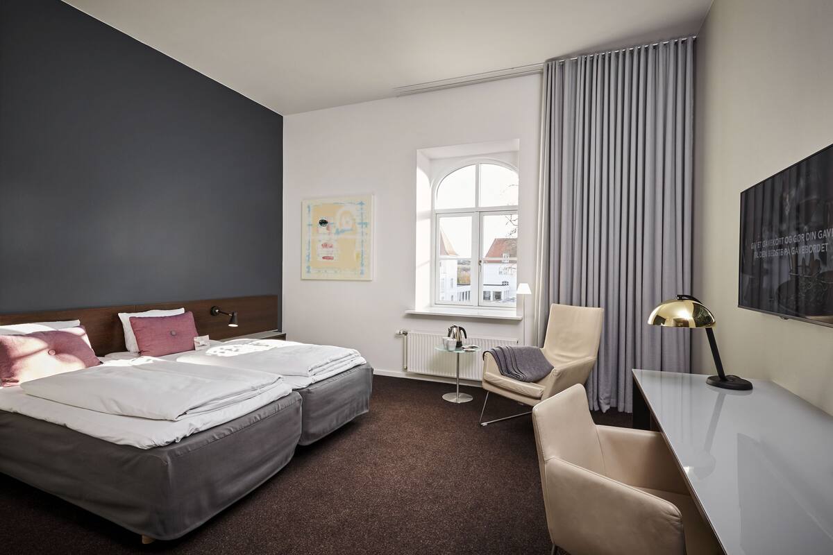 Habitación de hotel con dos camas, un escritorio, una silla, un ventanal con cortinas y un radiador.