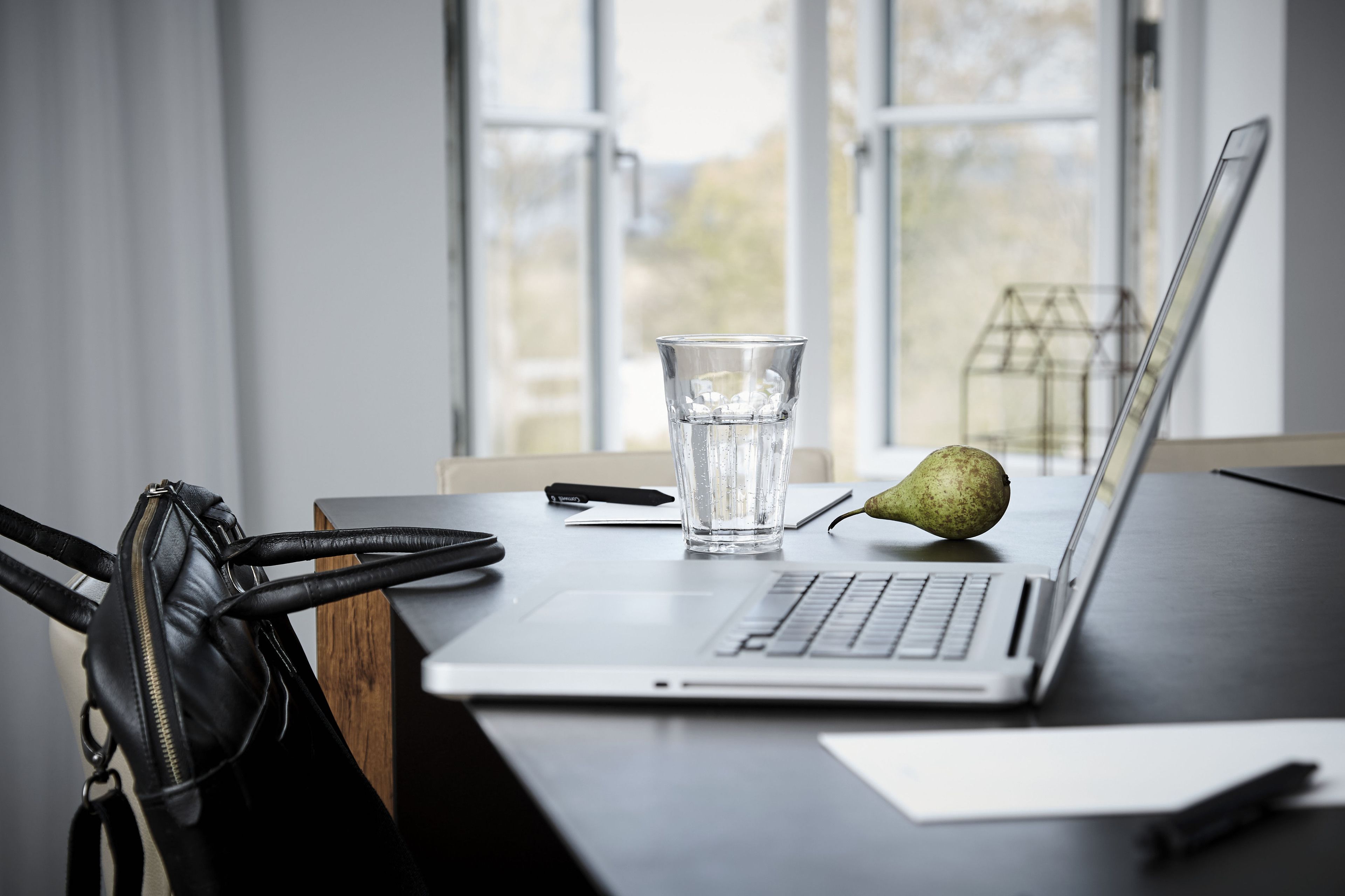 Ein Laptop auf einem Schreibtisch mit einem Glas Wasser, einem Birne und einer schwarzen Tasche.