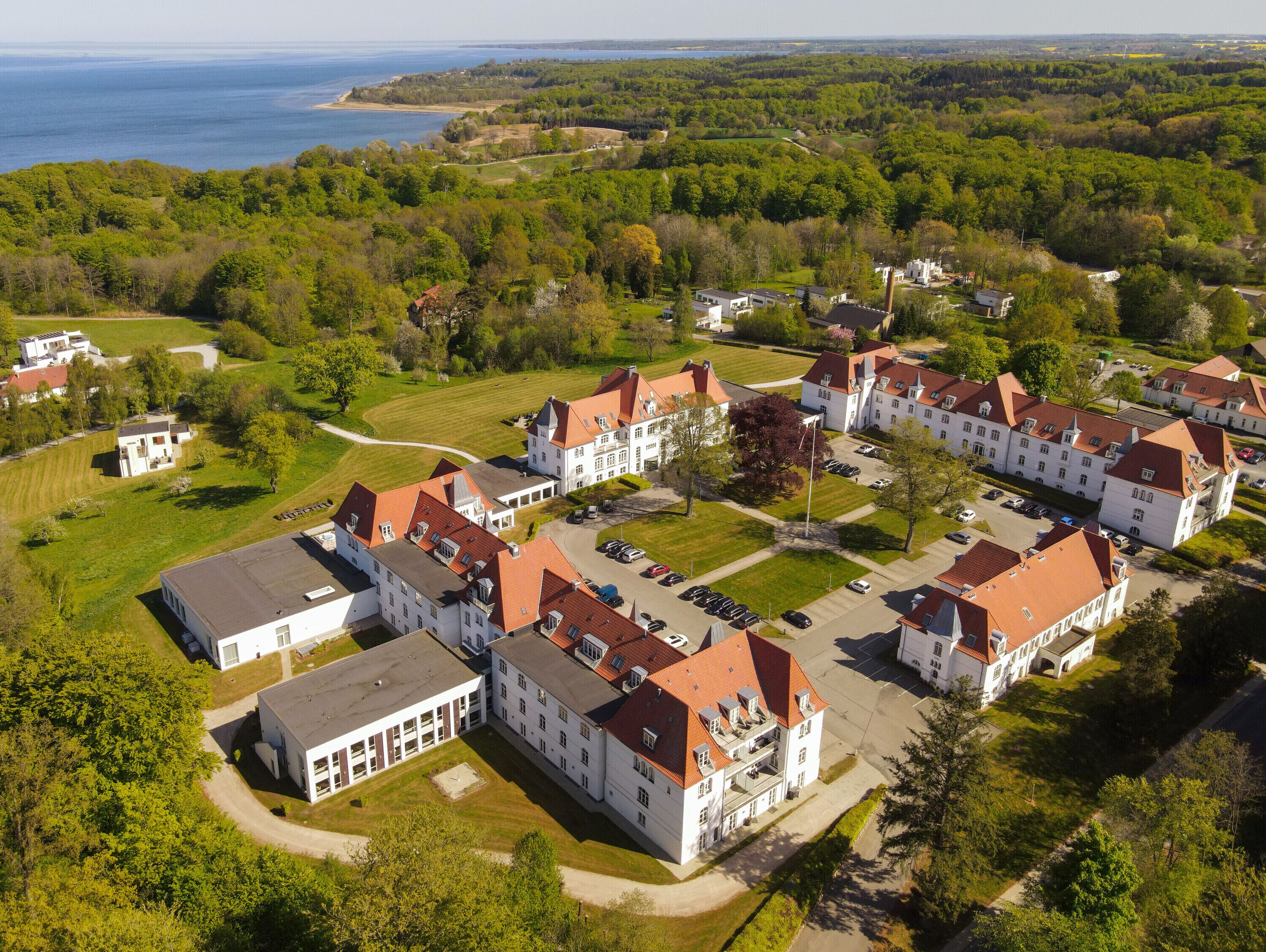 An aerial view of a large estate with multiple buildings, including a main house with a red roof and several smaller structures. There are well-maintained lawns and a body of water in the background.