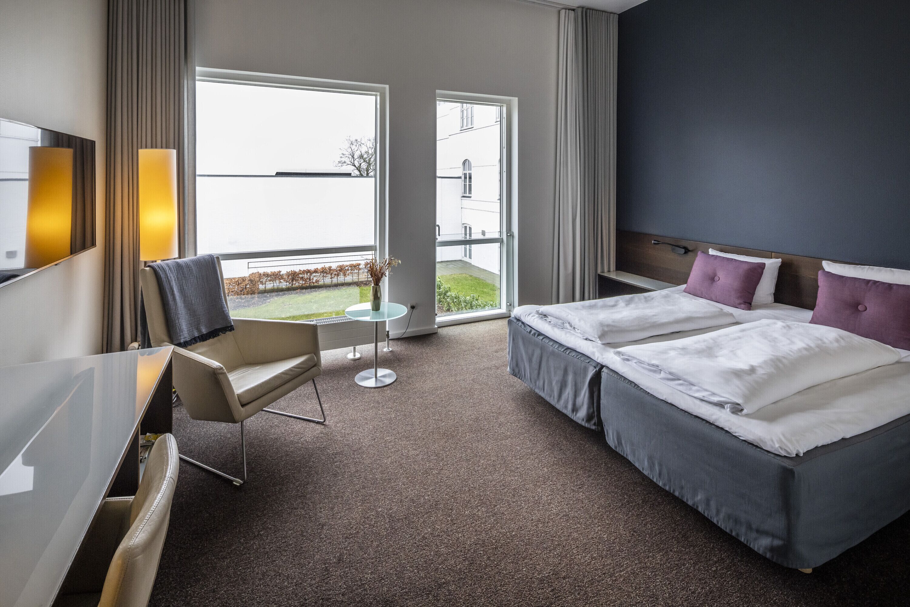 Ein Hotelzimmer mit zwei Betten, einem Schreibtisch, einem Sessel, einem kleinen Tisch und großen Fenstern.