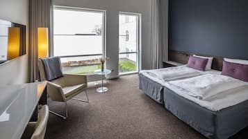 Ein Hotelzimmer mit zwei Betten, einem Schreibtisch, einem Sessel, einem kleinen Tisch und großen Fenstern.