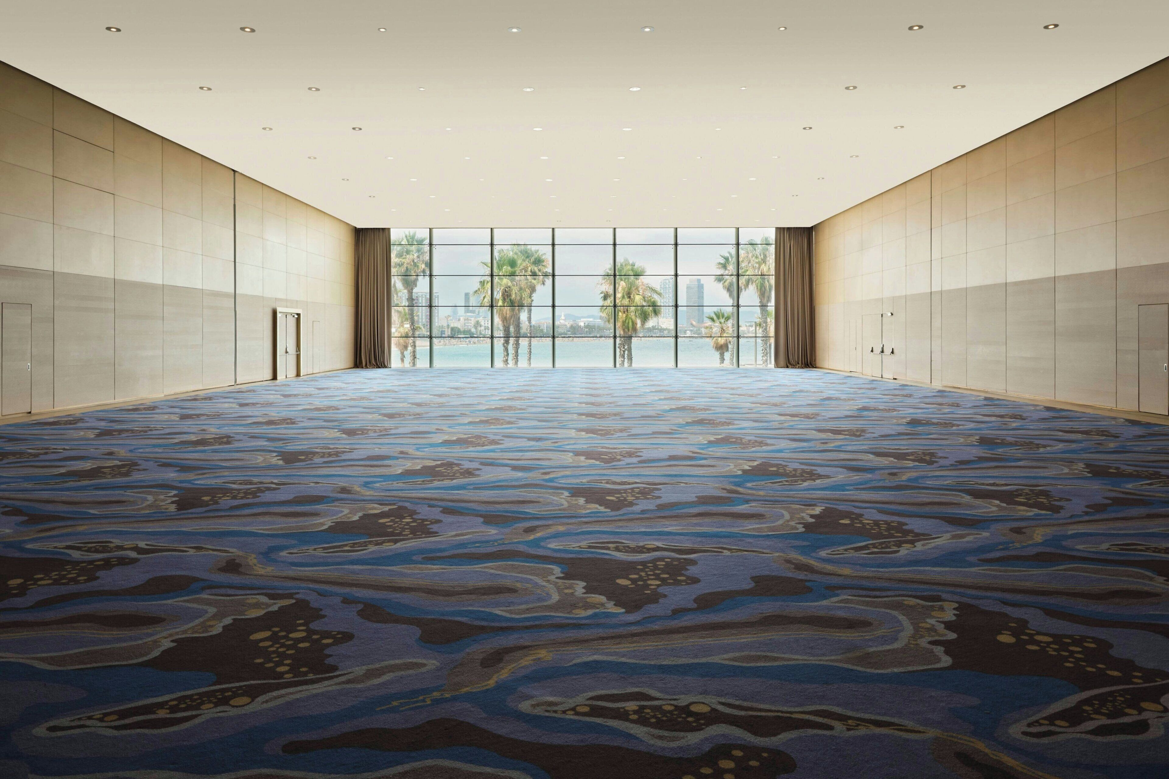 Un salón de conferencias amplio y vacío con alfombra estampada, ventanas grandes y vistas a palmeras y edificios.