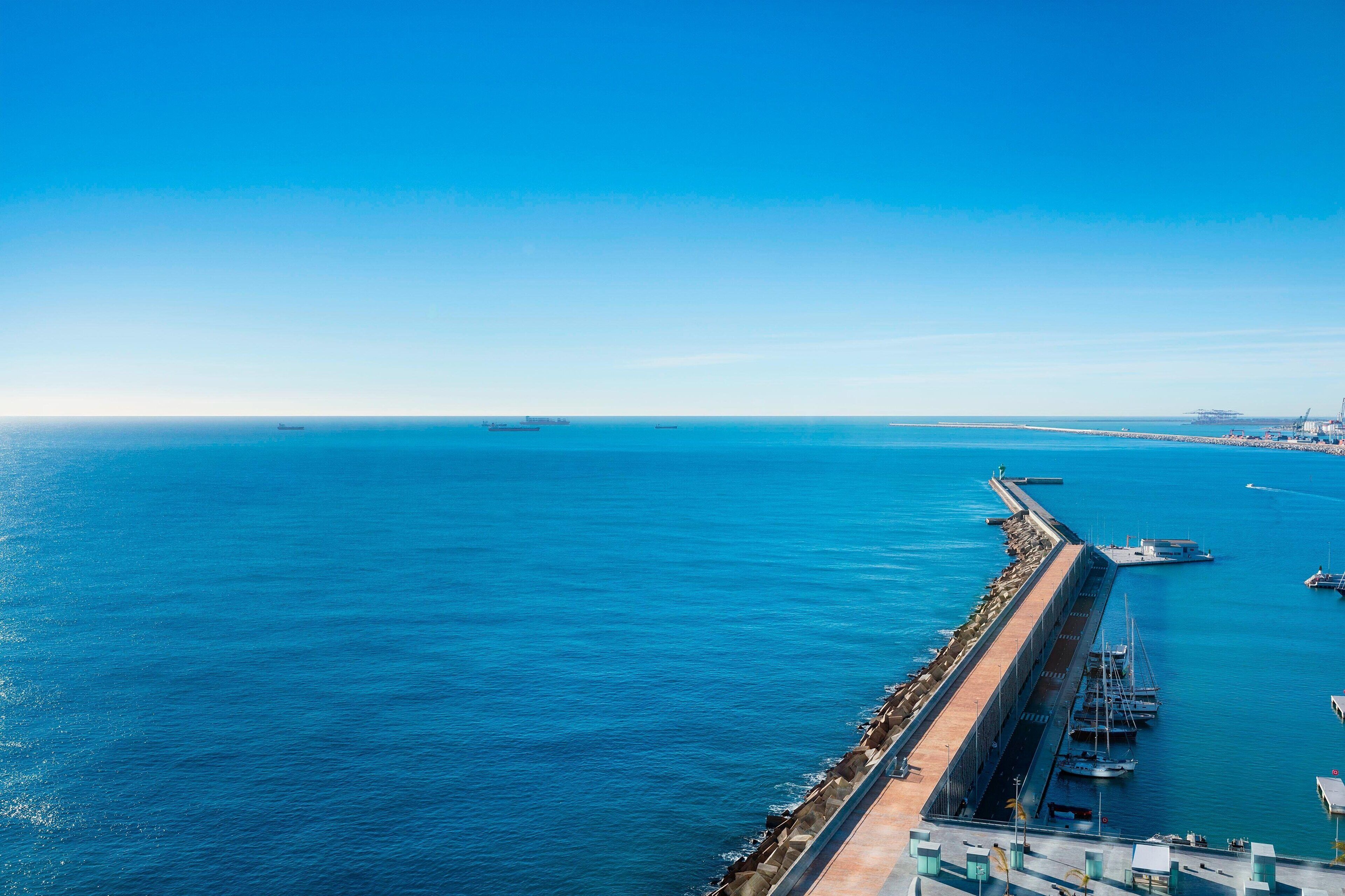 A coastal marina with a long pier extending into the water, several boats docked, and a clear blue sky.
