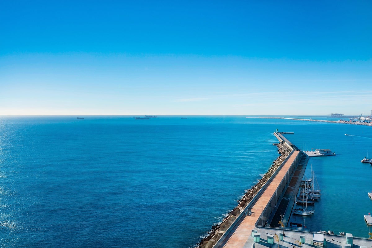 Un port de plaisance situé sur la côte, avec une longue jetée s’avançant dans l’eau, plusieurs bateaux amarrés et un ciel bleu dégagé.
