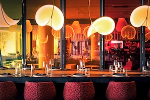 A restaurant with a long table set for dining, multiple hanging pendant lights, and red patterned chairs.