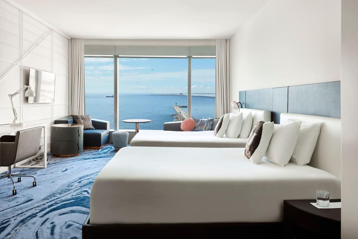 Quarto de hotel moderno com uma cama grande, uma escrivaninha, uma cadeira e vista para o mar.