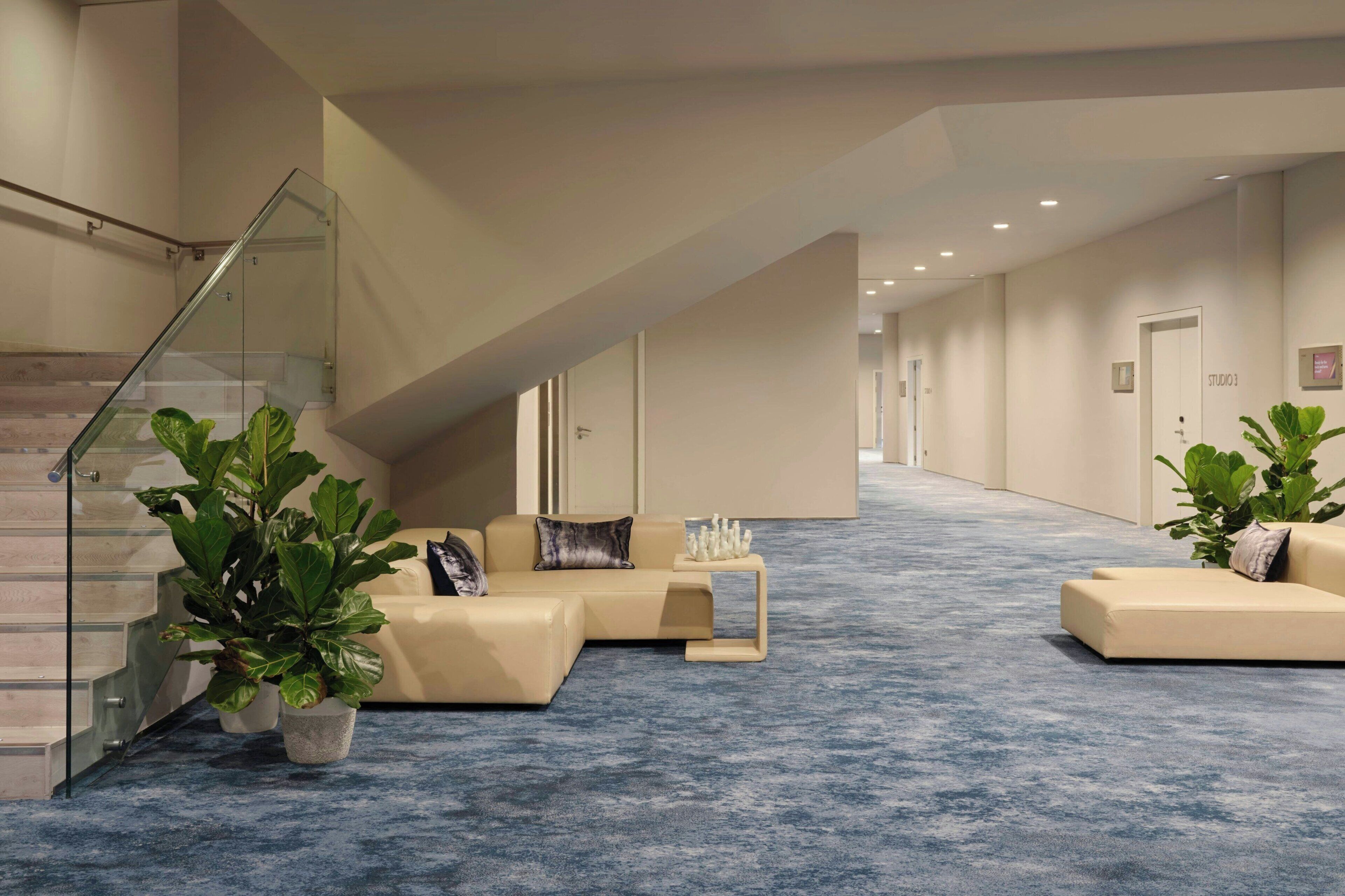 A modern hotel lobby with a blue carpet, a staircase, and two sofas with plants.