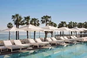 Un área junto a la piscina con sillas de descanso blancas y sombrillas, palmeras y vista al océano.