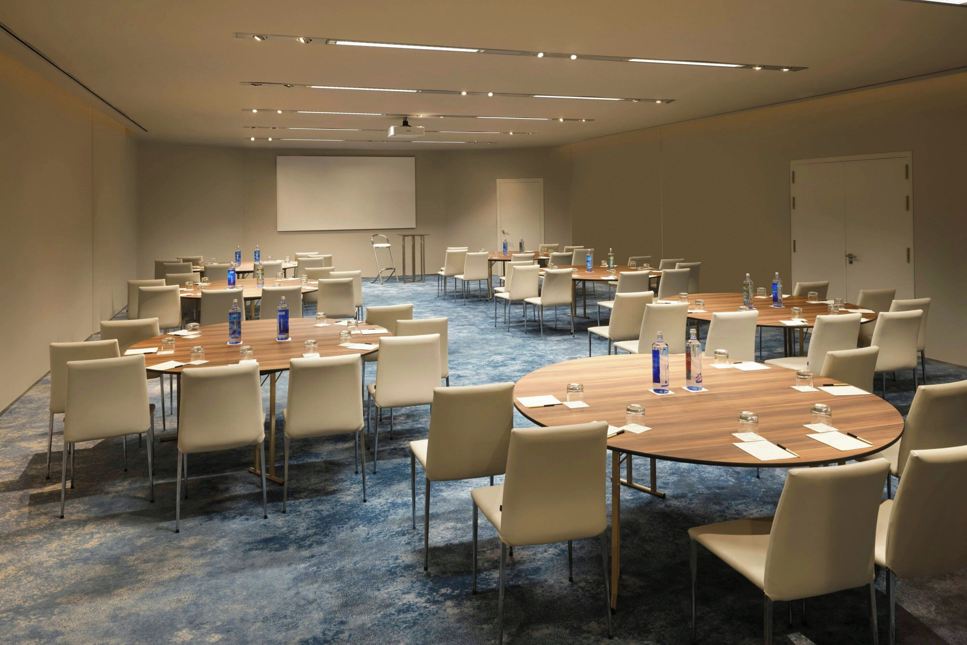 Un salón de conferencias con varias mesas y sillas, equipado con botellas de agua y vasos.
