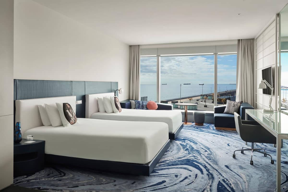 Quarto de hotel com duas camas, uma mesa, uma cadeira e vista para o mar.