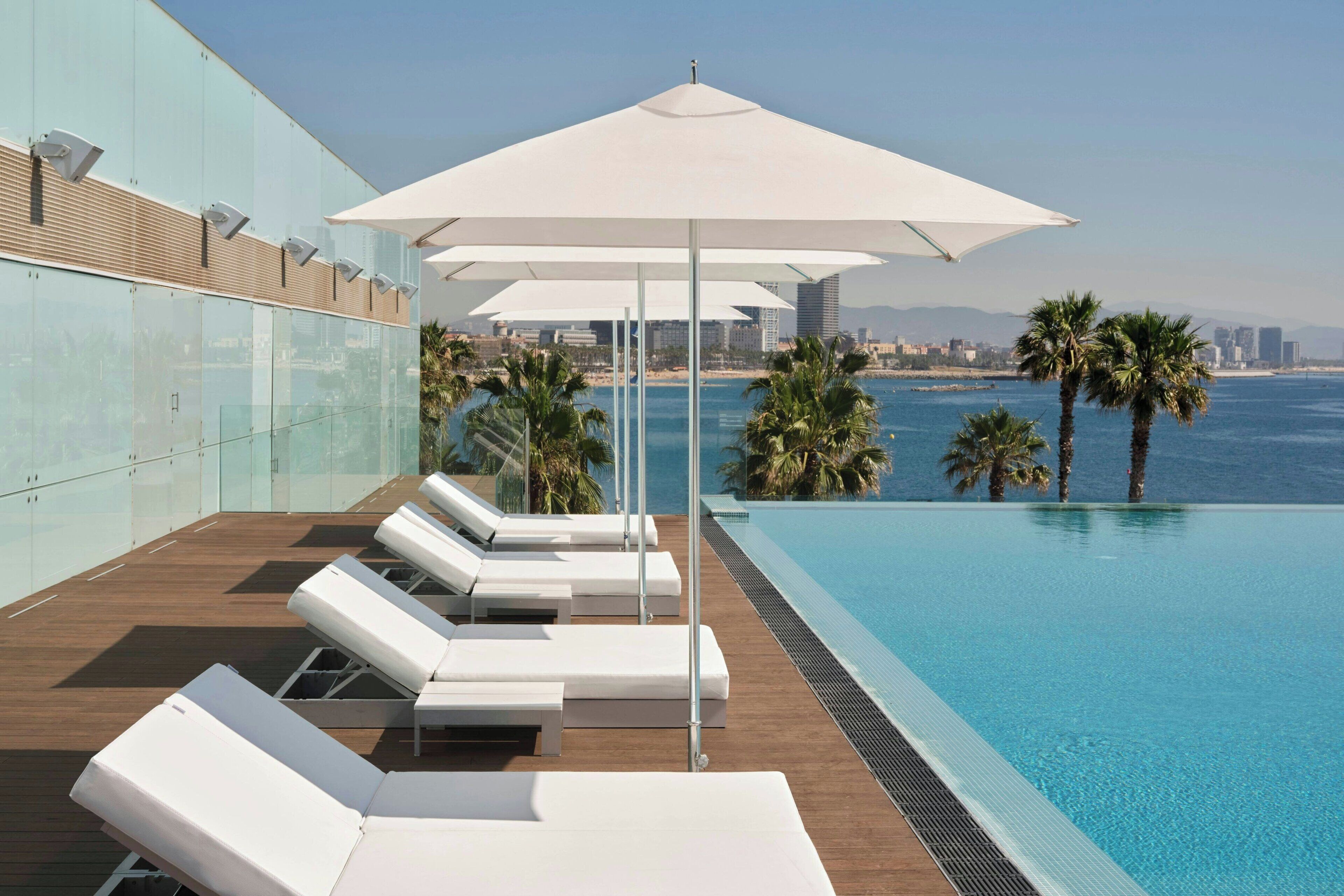Una piscina infinita en la azotea con sillas reclinables y sombrillas blancas, con vistas a la ciudad y al mar.