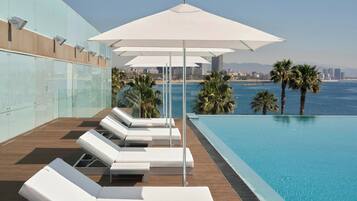 Una piscina infinita en la azotea con sillas reclinables y sombrillas blancas, con vistas a la ciudad y al mar.