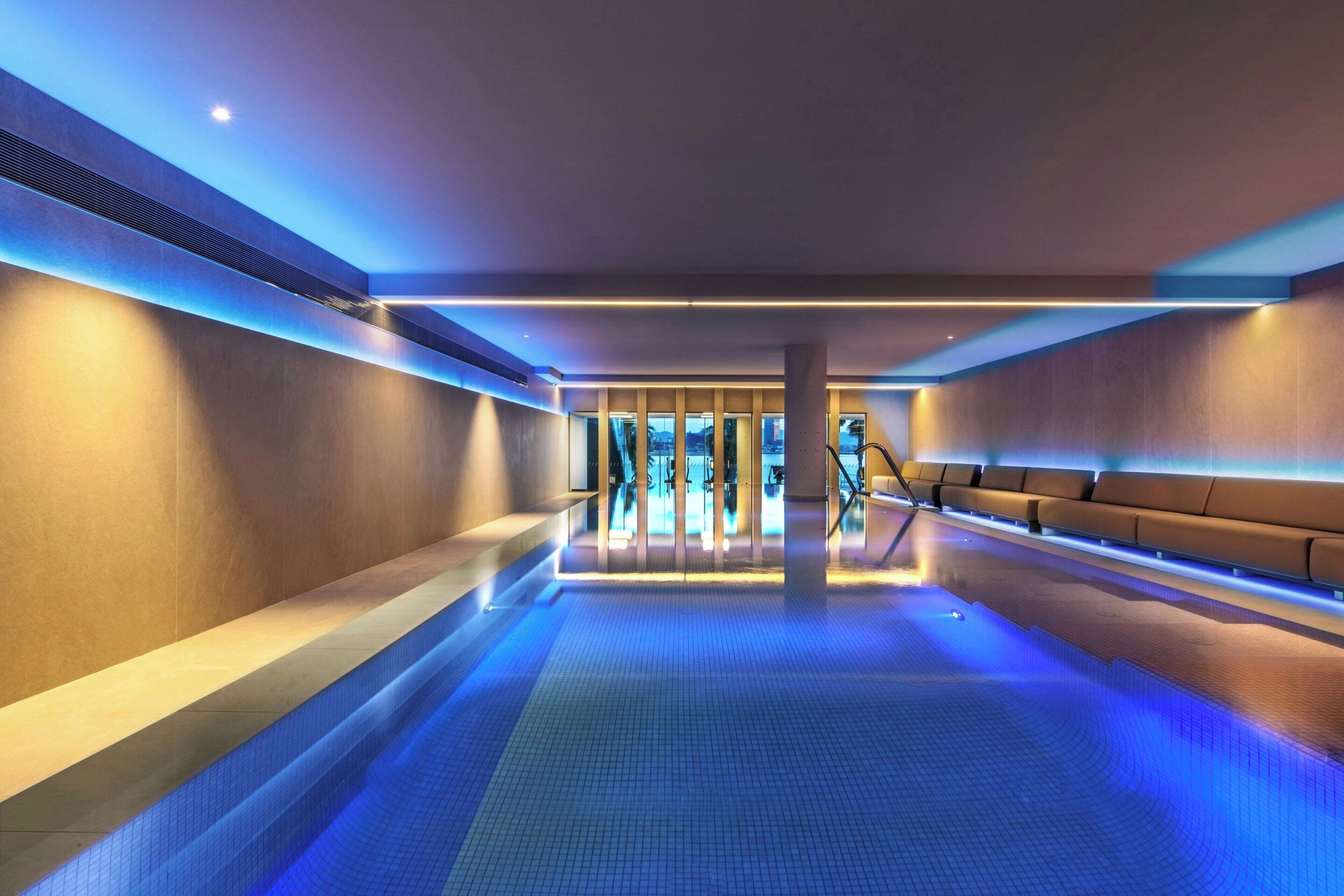 Piscina interior moderna con iluminación azul, una zona de estar y vistas al exterior.