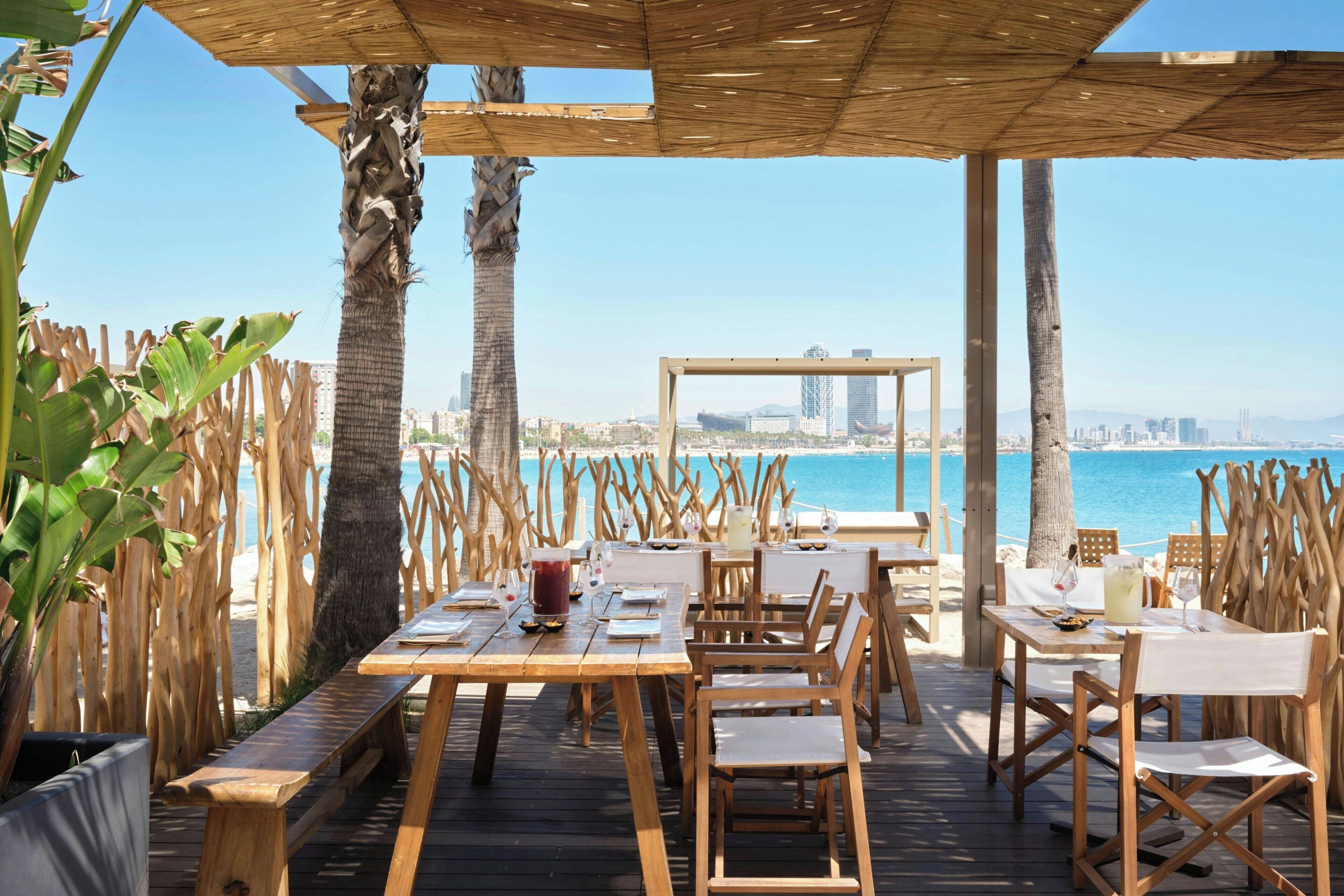 Een terras met houten tafels en stoelen, uitzicht op zee en een stadsachtergrond.