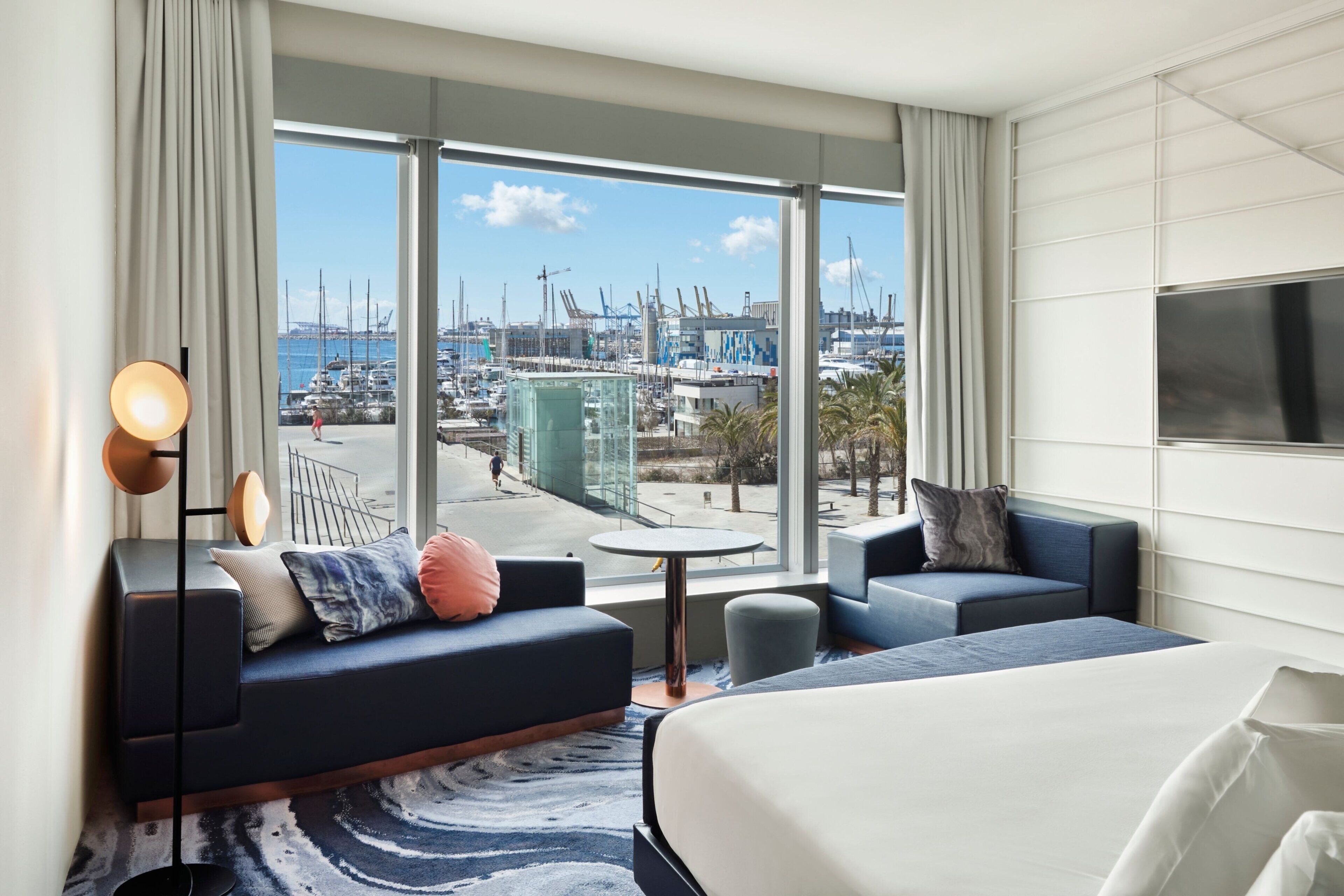 Habitación de hotel moderna con un gran ventanal que ofrece vista al puerto deportivo, un sofá, una cama y una pequeña mesa redonda.
