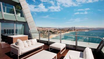 Un área de lounge al aire libre moderna con un sofá blanco, un sillón blanco y una mesita. Una barandilla de cristal da a la playa y al paisaje urbano.