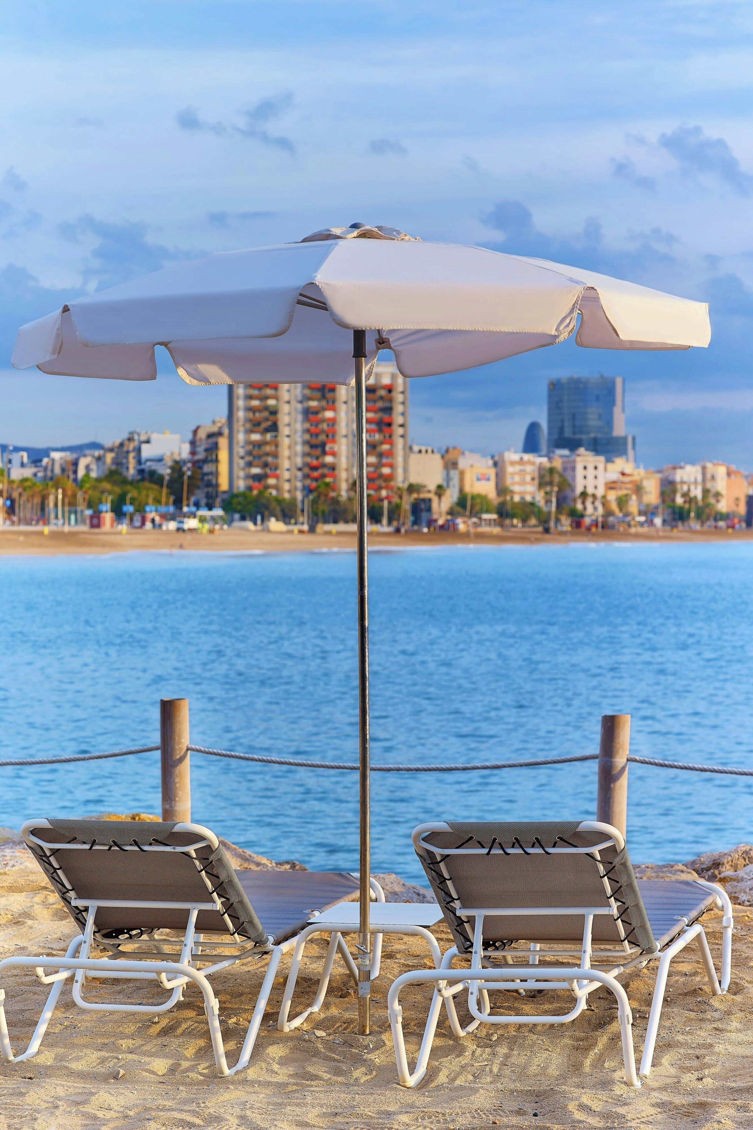 A beachside view with two lounge chairs and a white umbrella, overlooking a cityscape and the sea.