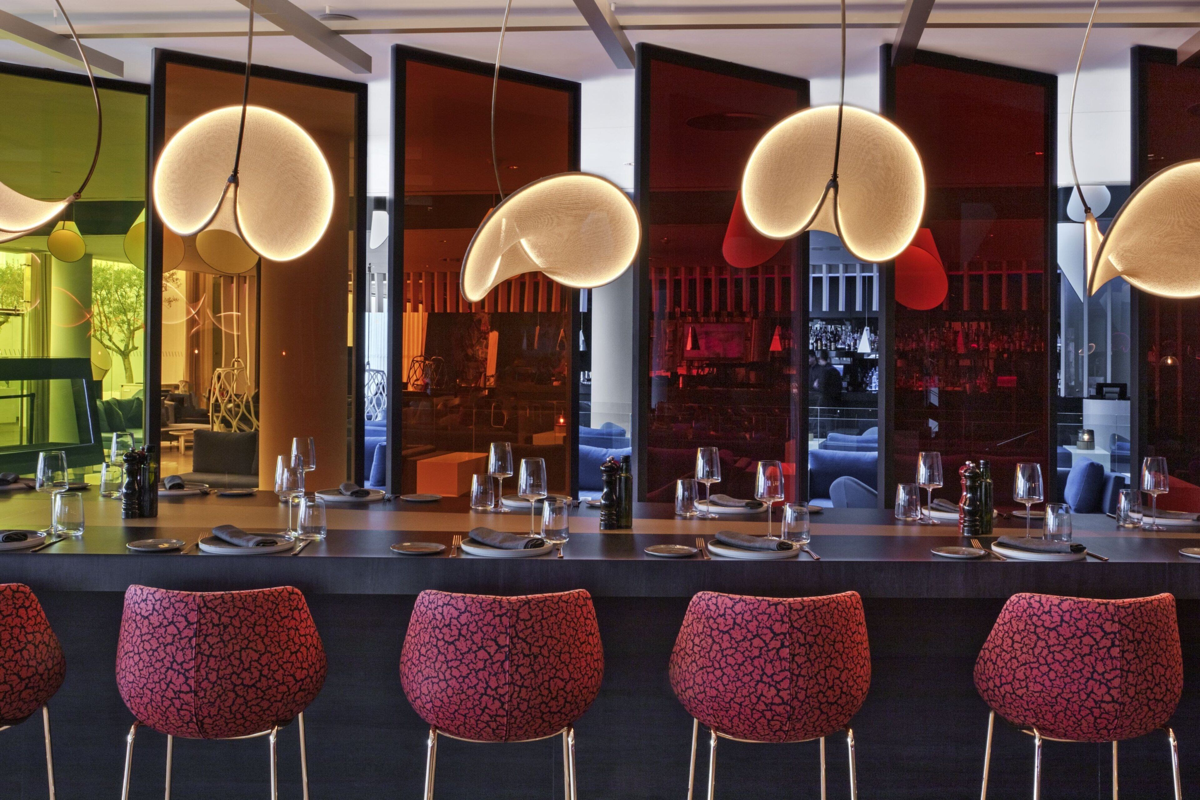 A modern bar area with red patterned chairs, a long counter, and pendant lighting.