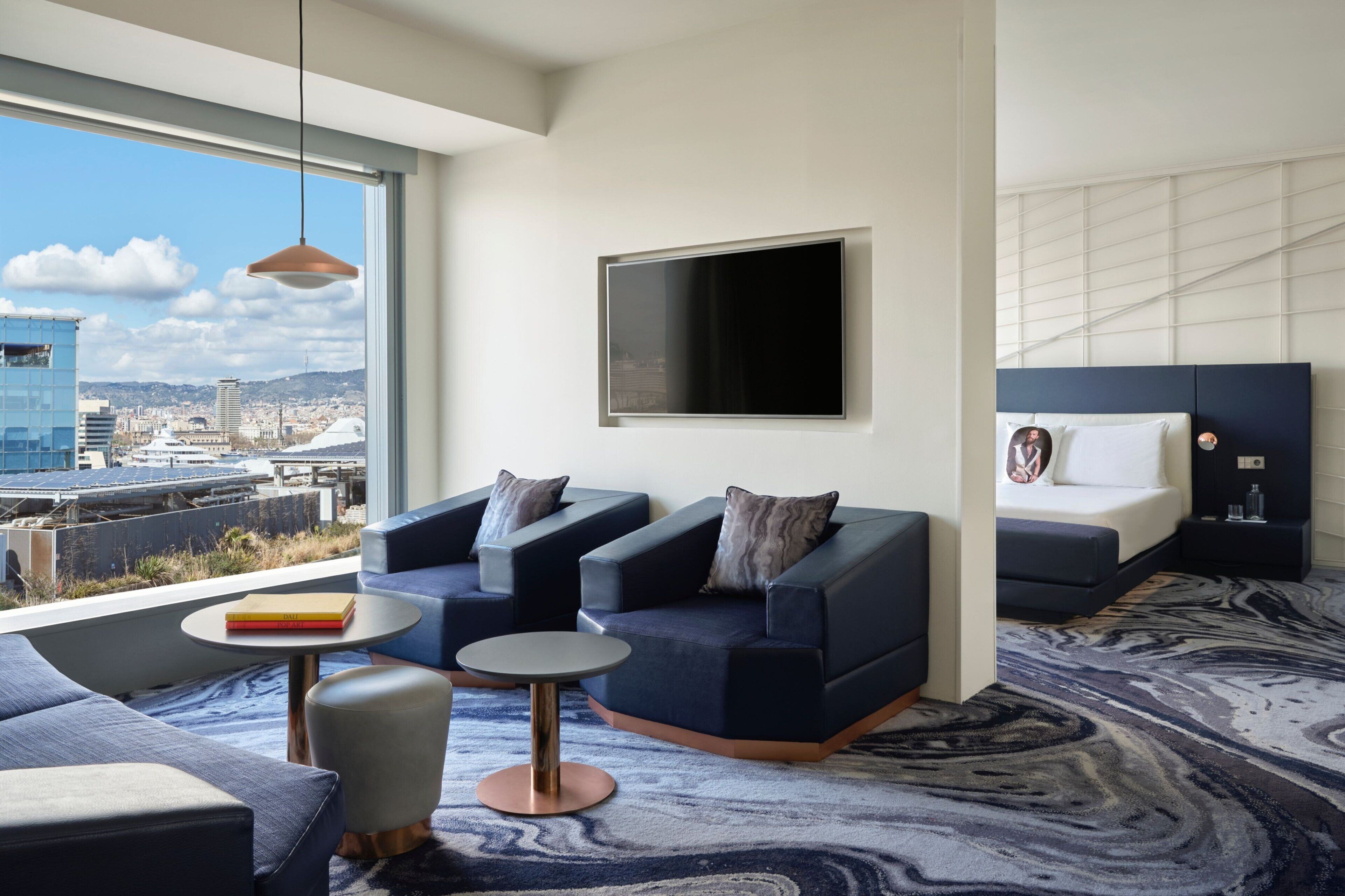 Une chambre d’hôtel moderne avec vue sur la ville, une grande télévision à écran plat, un canapé, une petite table ronde et un tapis à motifs.