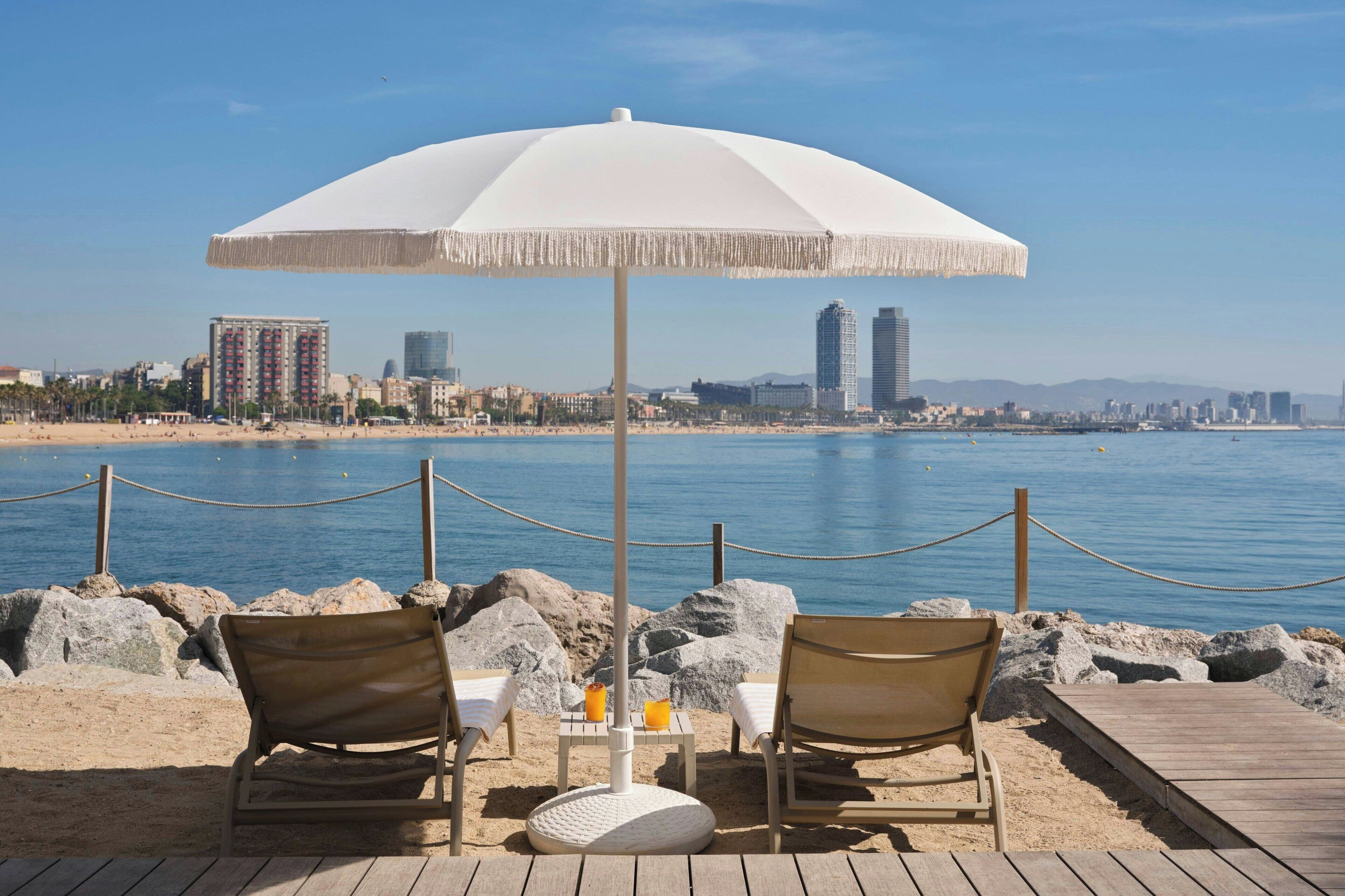 A beachside seating area with a white umbrella, two chairs, and a table with drinks, overlooking a city skyline and the sea.