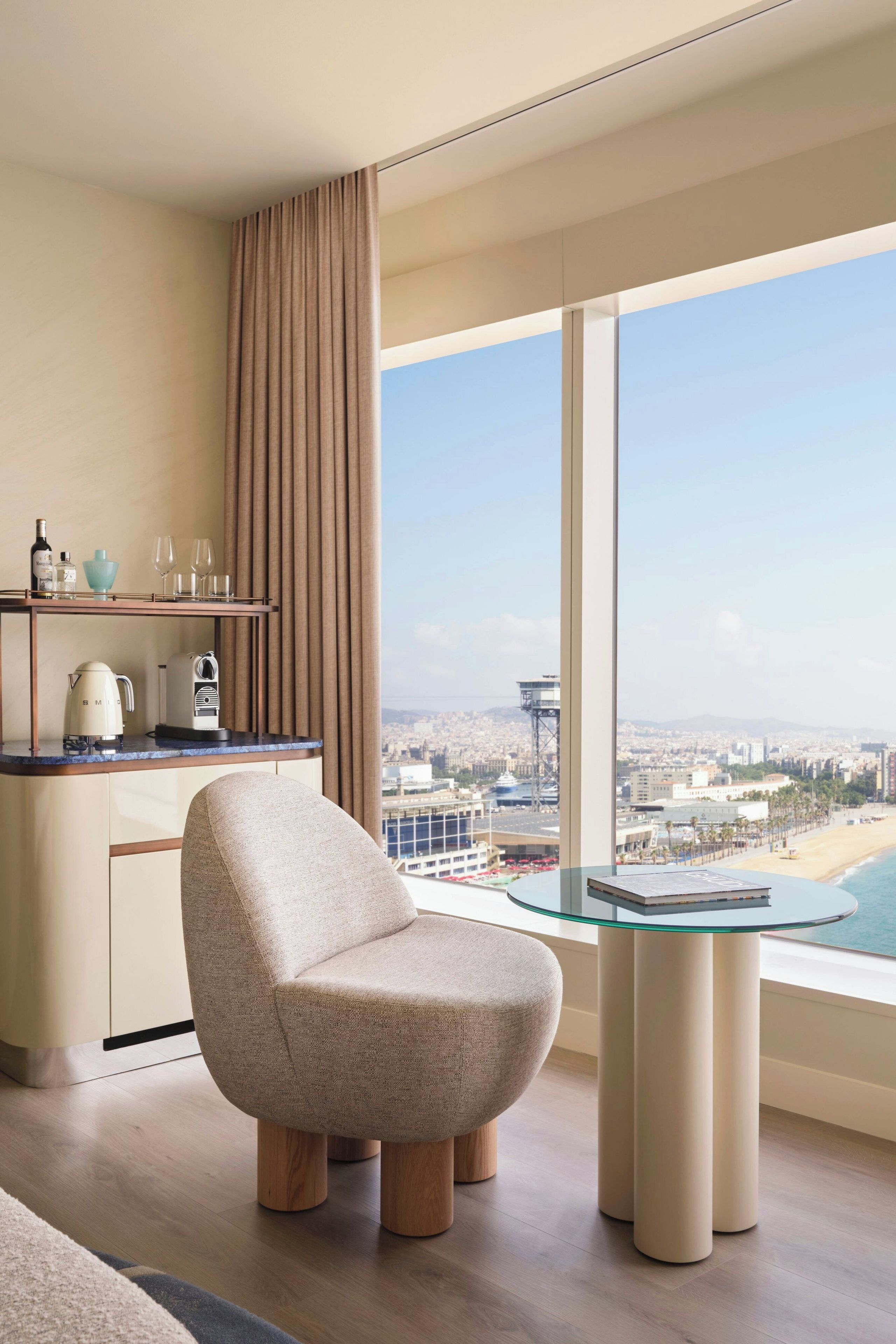 A modern hotel room with a city view, a round glass table, a cushioned armchair, and a minibar.