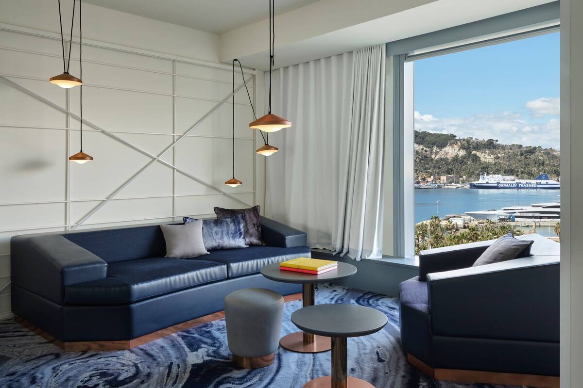 Uma sala de estar moderna com sofá, pufes e cadeiras, uma mesa de centro redonda, luminárias pendentes e uma grande janela com vista para um porto e colinas.