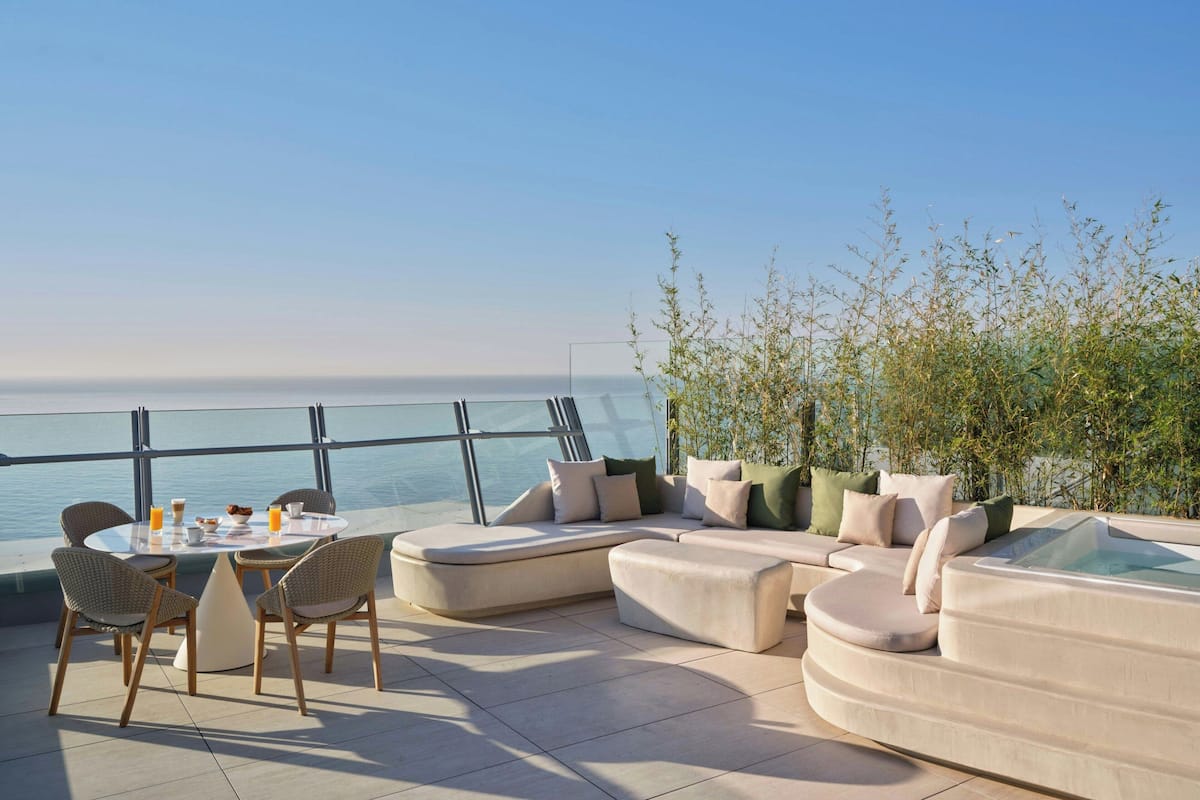 Um terraço com mesa de jantar, cadeiras, um sofá e uma banheira de hidromassagem, com vista para o mar.