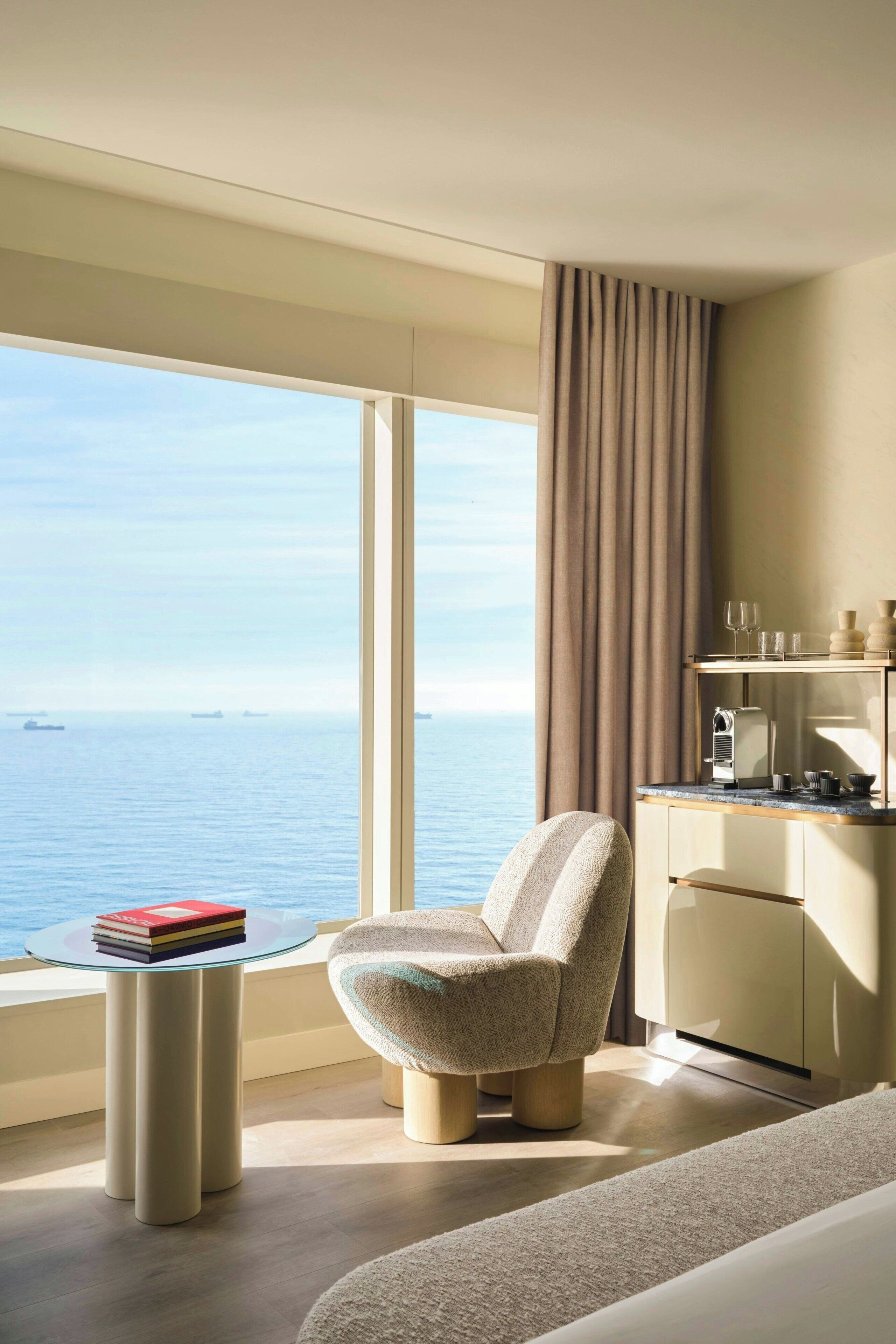 A modern hotel room with a large window, a round glass table, a cushioned chair, a minibar, and a bed.