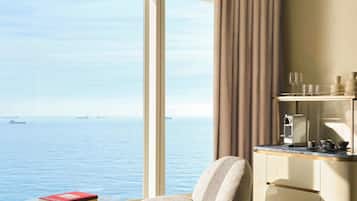 A modern hotel room with a large window, a round glass table, a cushioned chair, a minibar, and a bed.