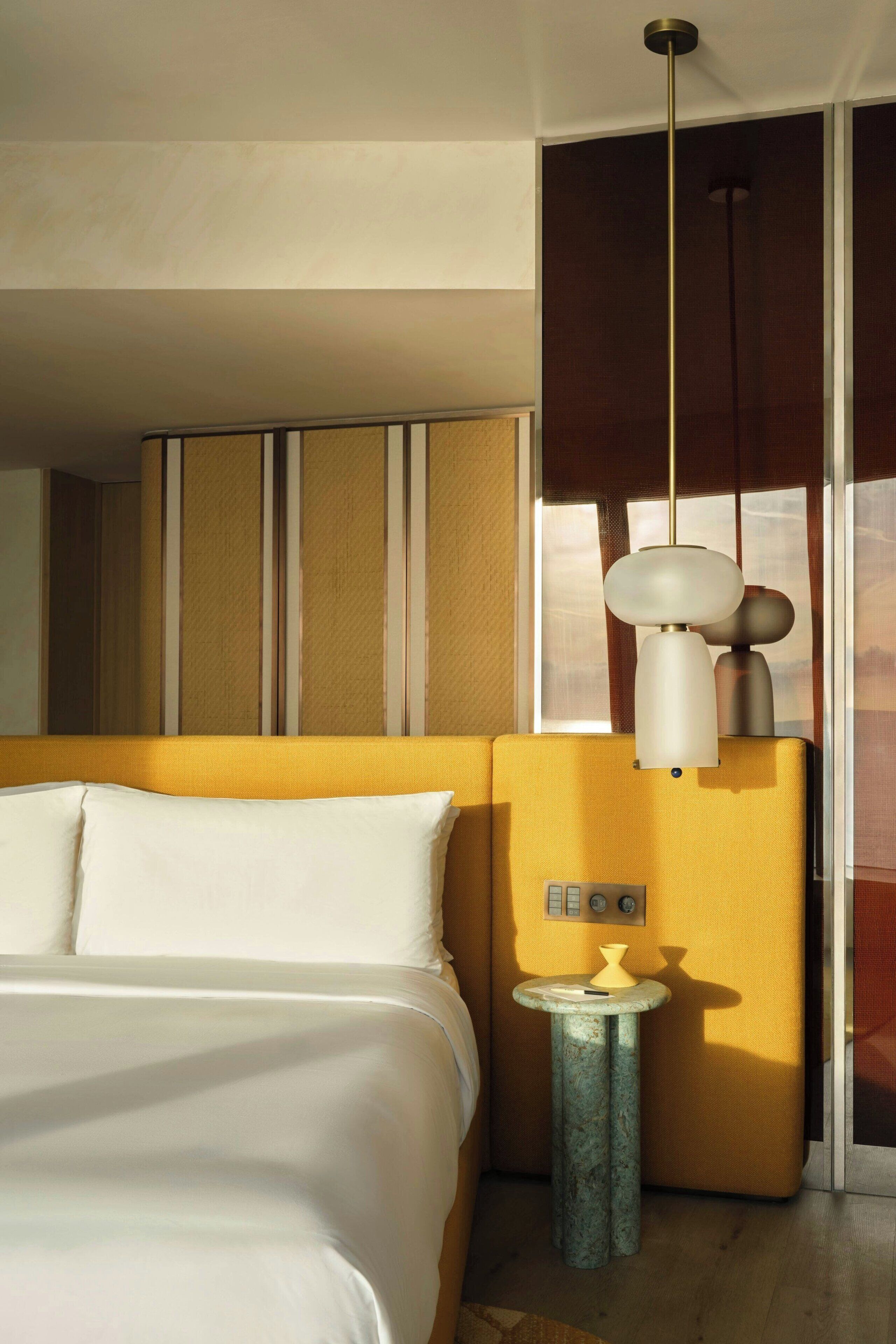 Habitación de hotel con una cama, una mesita de noche con lámpara, un lavamanos pequeño y una ventana con persianas.