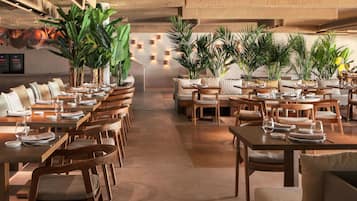 En rymlig restaurang med långa bord och stolar, omgiven av stora krukväxter.