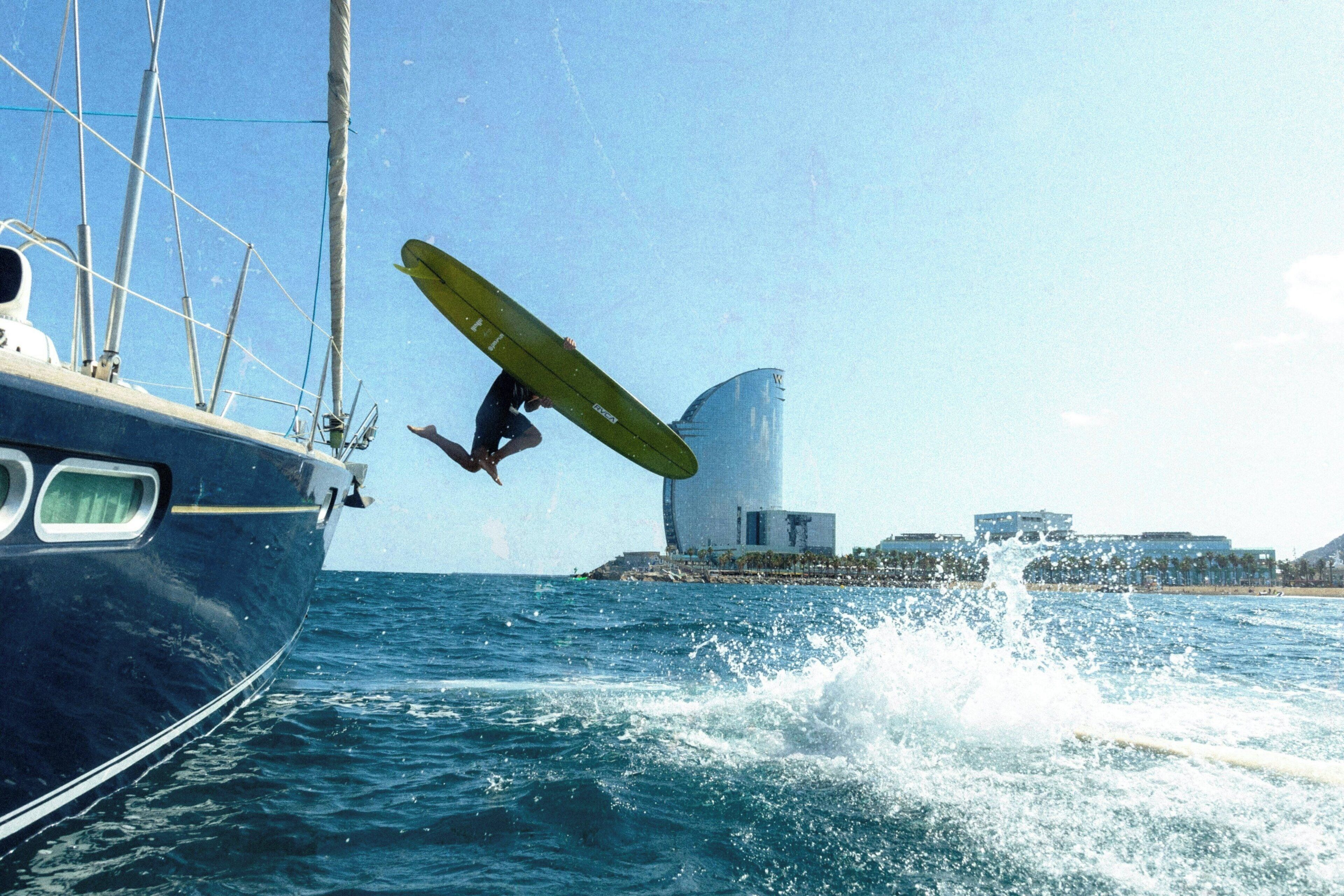 Una persona esegue un'acrobazia su una tavola da surf nell'oceano, con una barca a vela e degli edifici sullo sfondo.