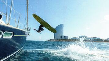 Una persona esegue un'acrobazia su una tavola da surf nell'oceano, con una barca a vela e degli edifici sullo sfondo.