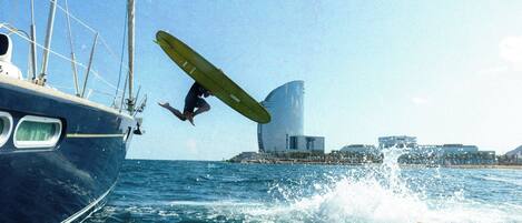 A person performing a stunt on a surfboard in the ocean, with a sailboat and buildings in the background.