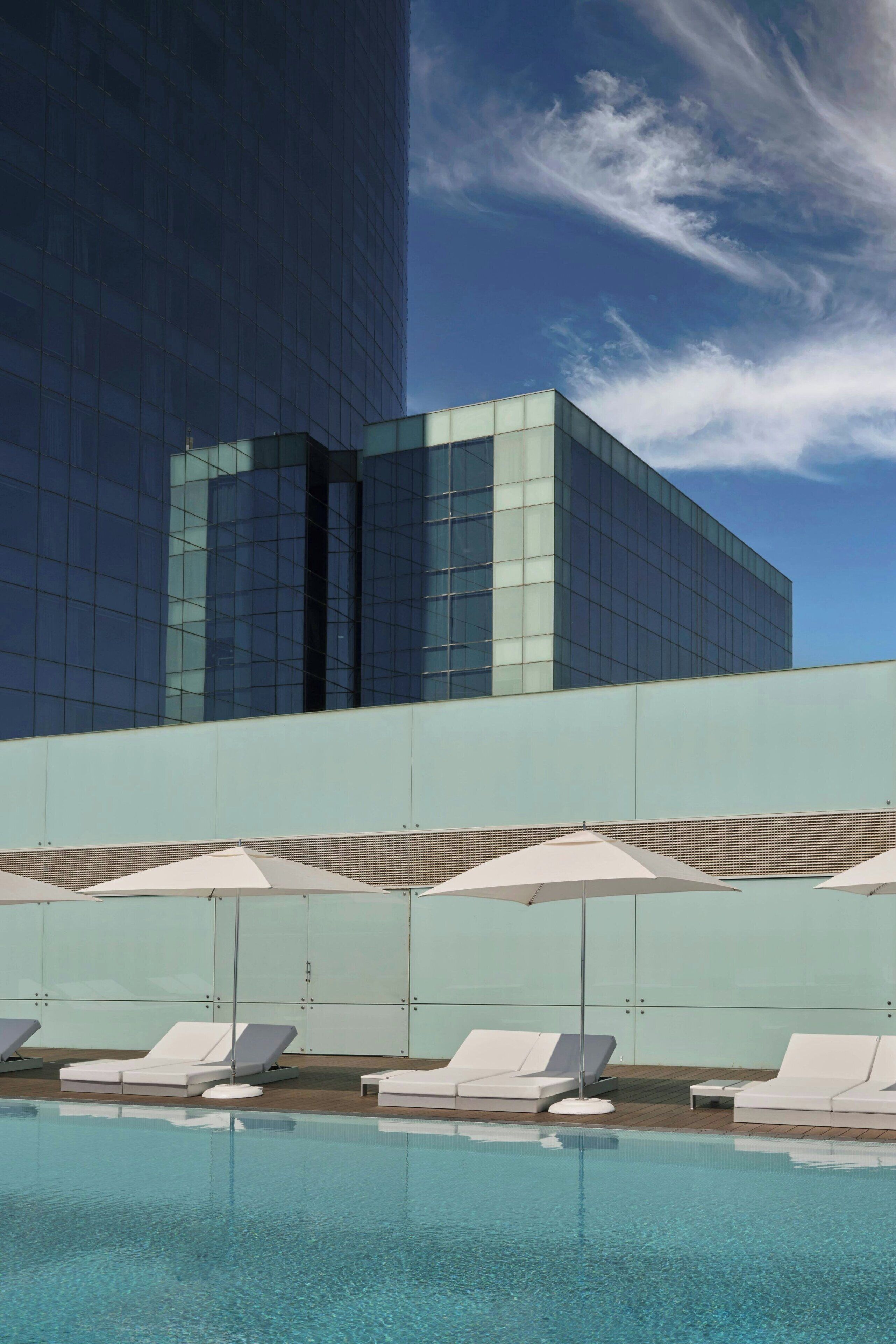 Un edificio moderno con fachada de vidrio, un área de piscina en la azotea con tumbonas y sombrillas, y un cielo despejado con algunas nubes.