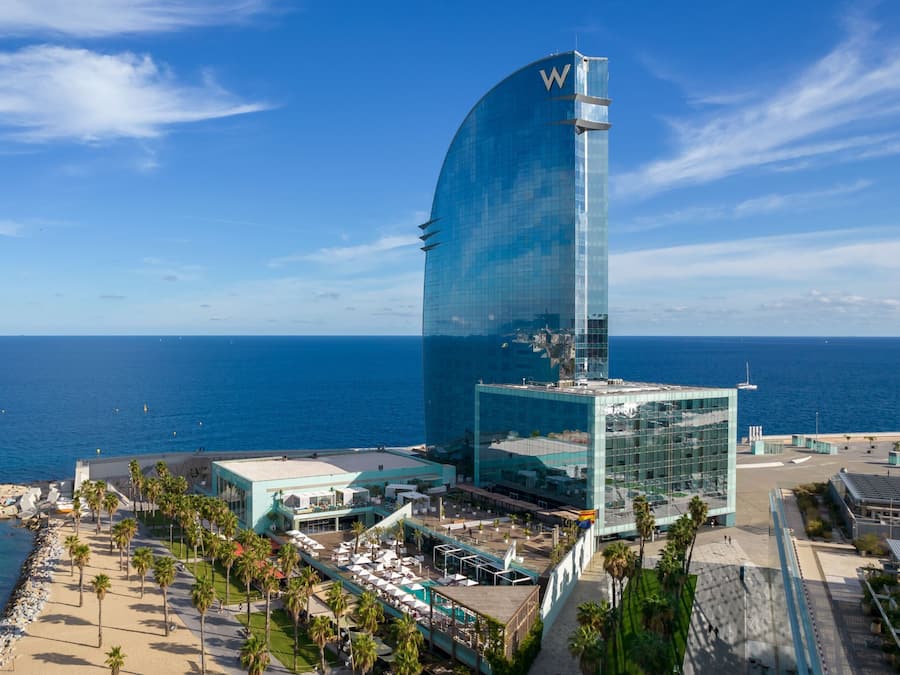 Un hotel moderno con fachada de vidrio curvada, situado cerca de una playa con palmeras y vistas al océano.