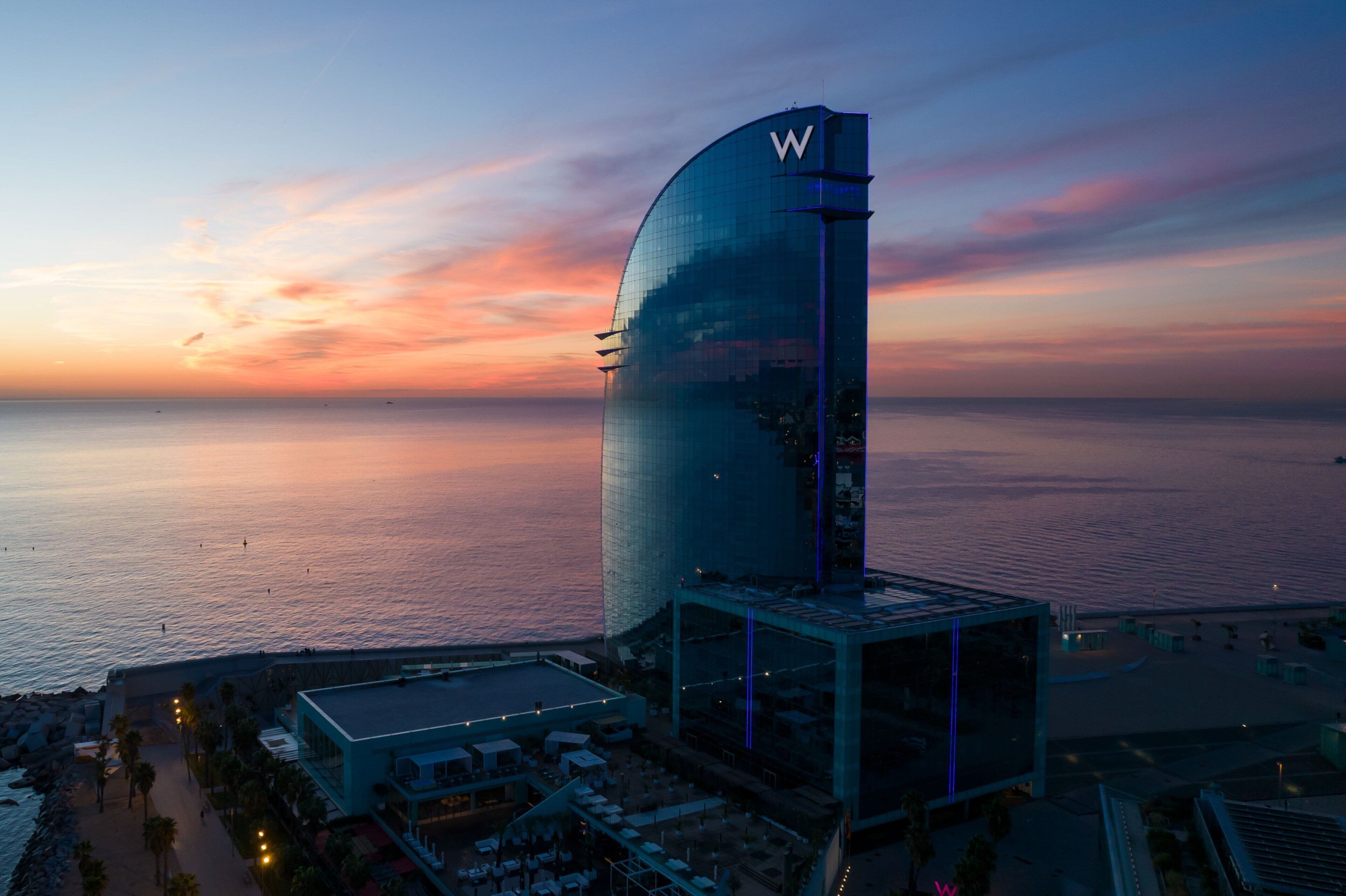 A modern hotel with a curved glass facade, a prominent 'W' logo, and a view of the sea at sunset.