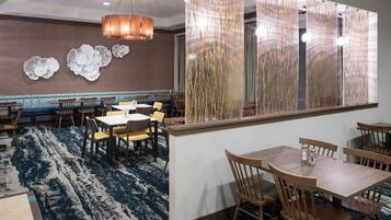 Restaurante com carpete azul estampado, mesas e cadeiras de madeira, arte decorativa na parede e luminária pendente.