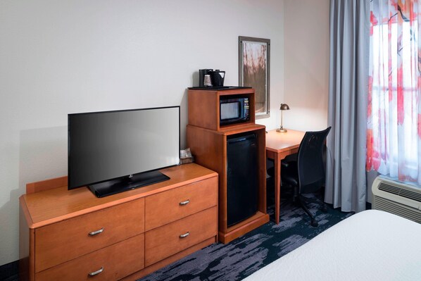 A hotel room with a bed, a TV on a wooden dresser, a microwave, a small desk with a chair, and a window with curtains.