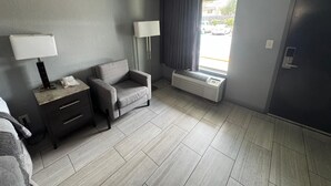 A hotel room with a grey sofa, a bedside table with a lamp, a window with curtains, and a door. - Budget Inn Sanford (Sanford)