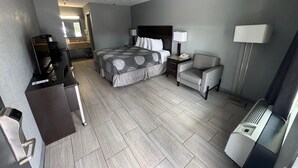 A hotel room with a bed, a desk, a chair, a nightstand, a lamp, and a heater. - Budget Inn Sanford (Sanford)