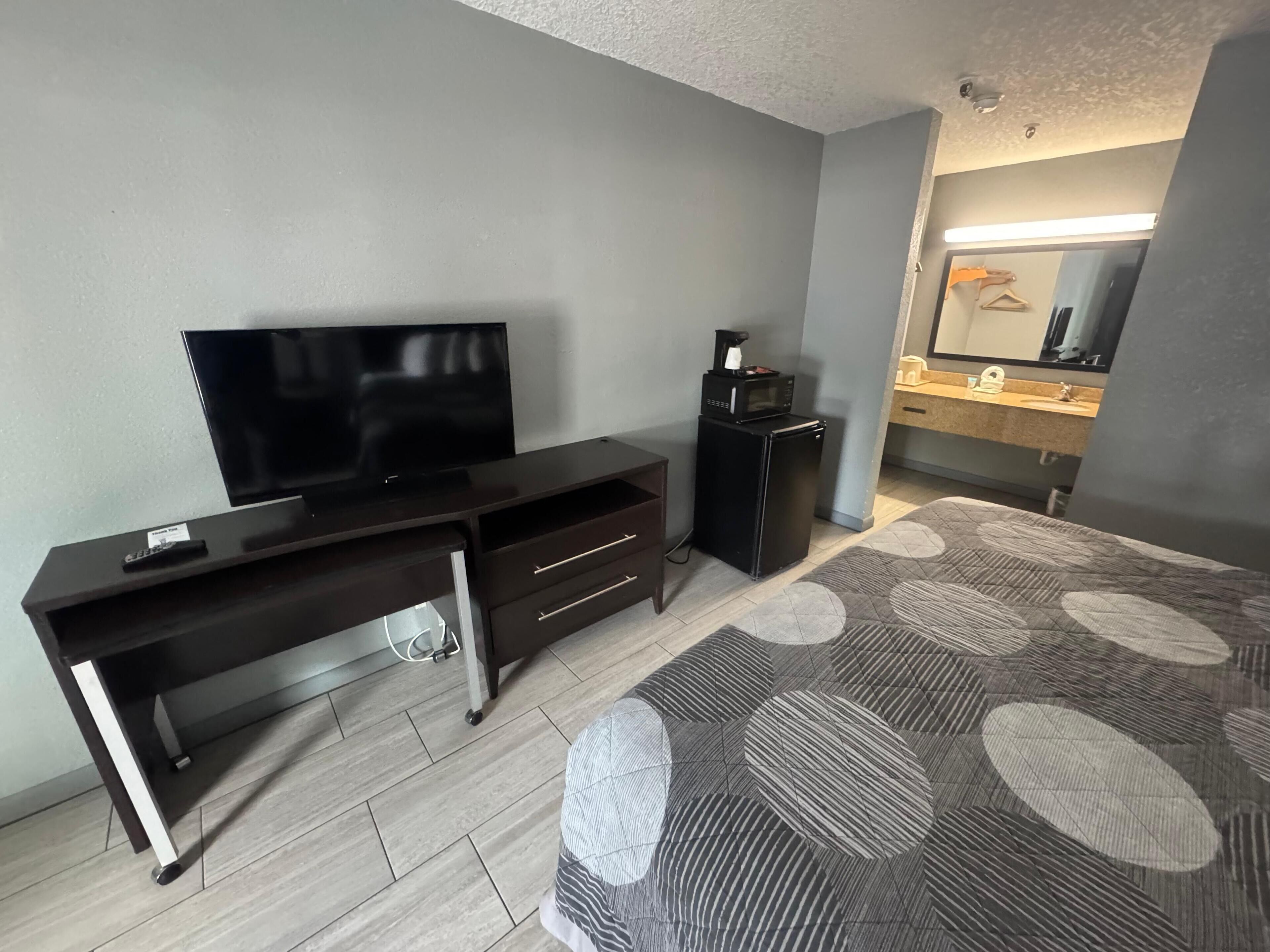 A hotel room with a flat-screen TV, a small black refrigerator, a desk with drawers, a patterned rug, and a bathroom with a sink and mirror.