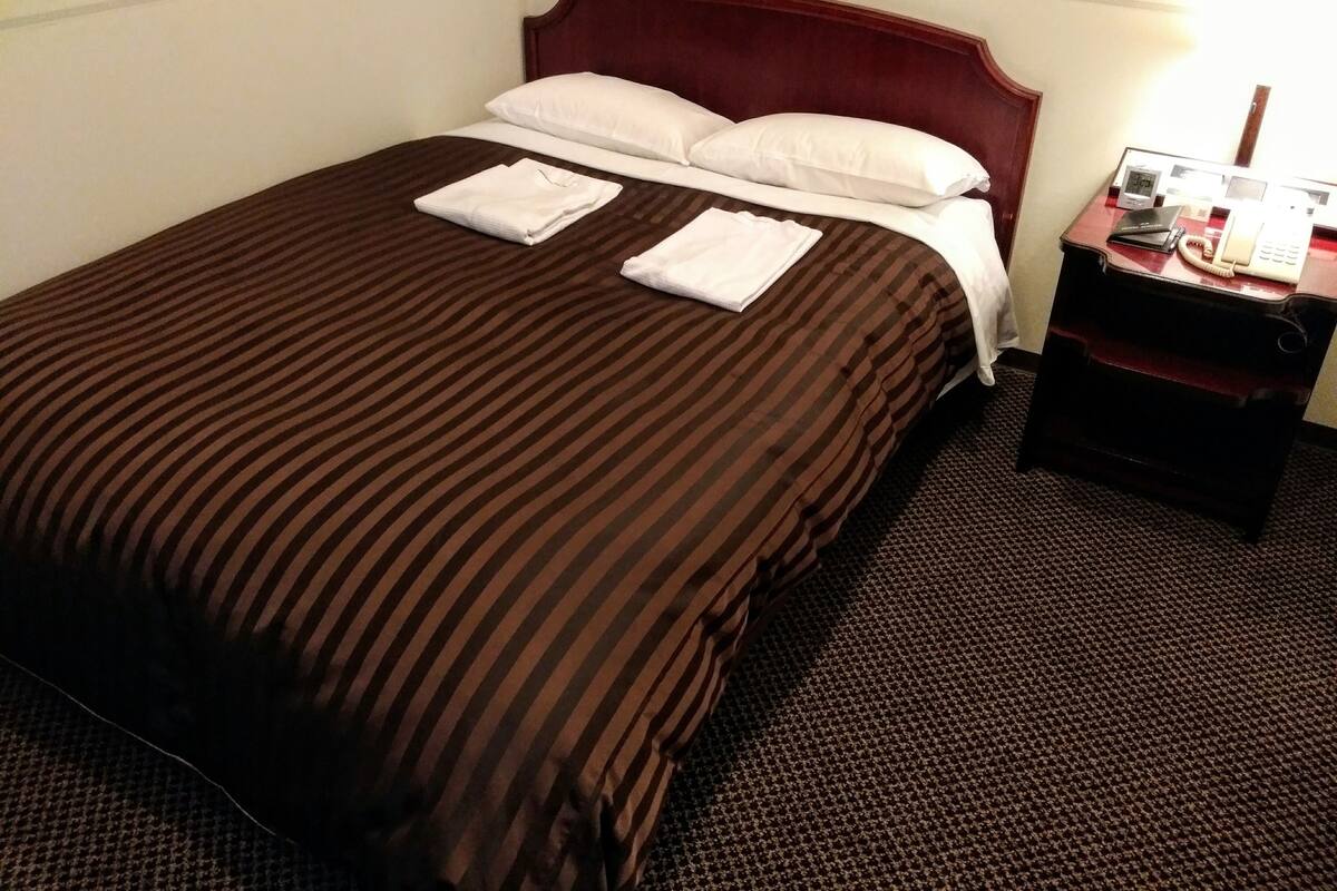 A hotel room with a large bed, two pillows, a neatly folded white towel, a wooden headboard, a nightstand with a telephone, and a carpeted floor.
