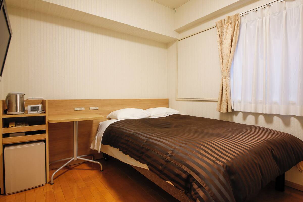 A bedroom with a bed, a small desk with a kettle, and a mini fridge.