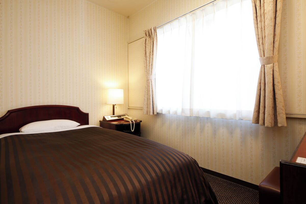 A hotel room with a large bed, a bedside table with a lamp, a telephone, and a window with curtains.