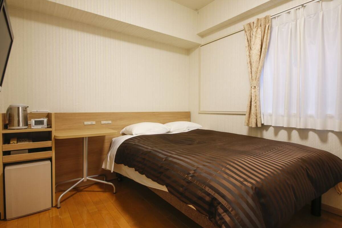 A bedroom with a bed, a desk, a small refrigerator, and a window with curtains.
