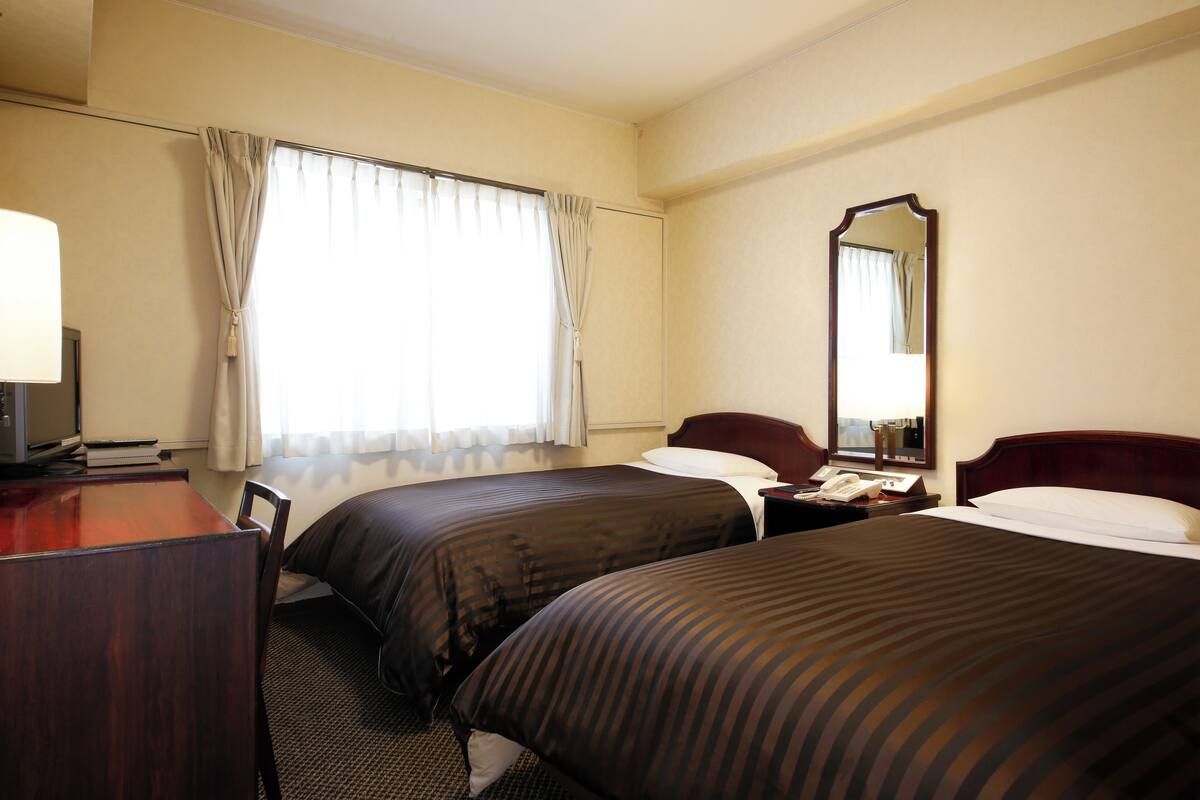 A hotel room with two beds, a desk with a lamp, a mirror, and a window with curtains.