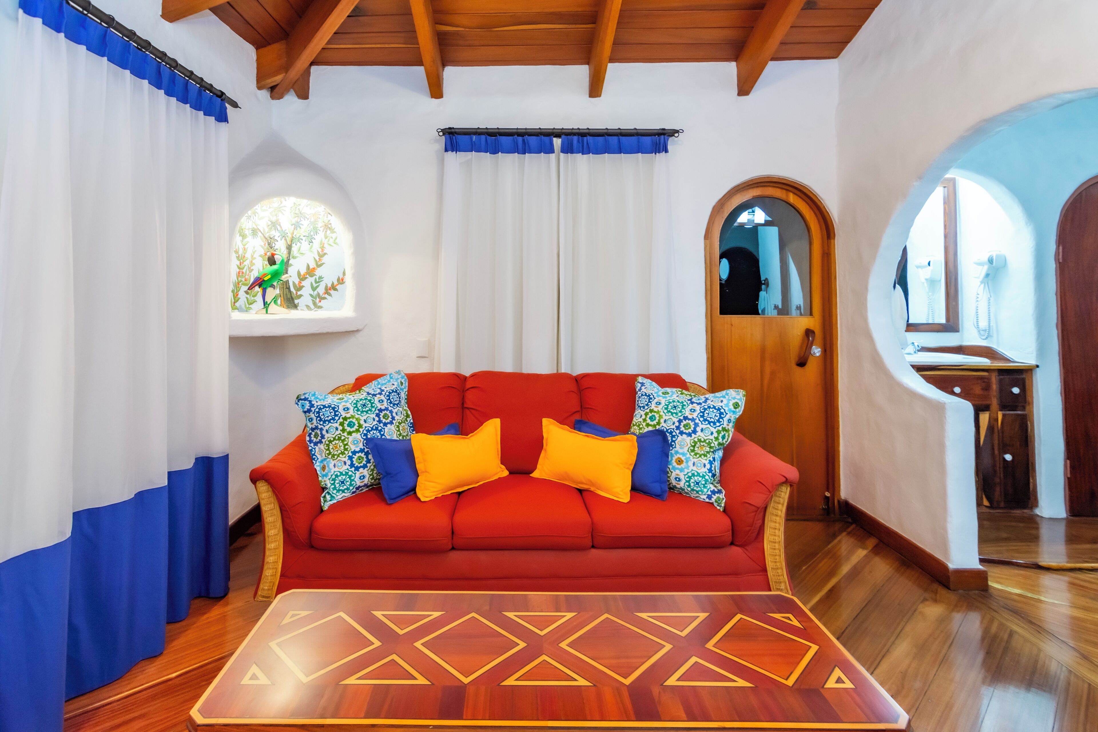 A red sofa with colorful cushions, a wooden coffee table, and a window with a stained glass view.