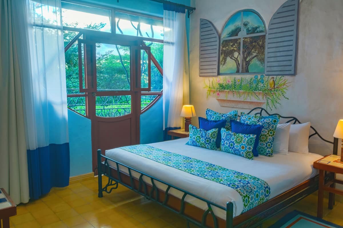 A bedroom with a bed, curtains, a window, a nightstand, and a wall-mounted shelf with plants.